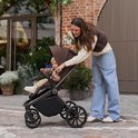 Een vrouw met een bruine kinderwagen met een baby erin, met een bos bloemen in haar hand.
