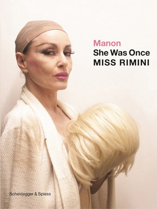 Vrouw met beige haarkapje houdt blonde pruik vast, met tekst 'Manon She Was Once MISS RIMINI' op witte achtergrond.