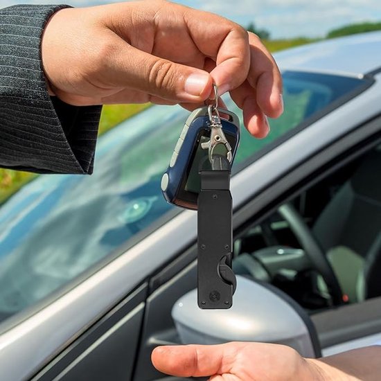 Een hand overhandigt een zwarte sleutelorganizer met autosleutels en een D-ring aan een andere hand, met een auto op de achtergrond.