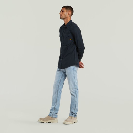 Man met donkerblauw G-Star overhemd en lichtblauwe jeans, staand met handen achter de rug.