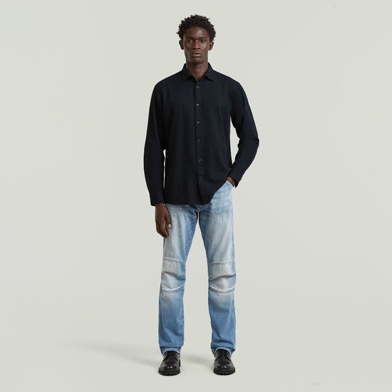 Man, hele lichaam, staat met de hand in de zak, draagt een donkerblauw regular fit overhemd en lichte jeans.