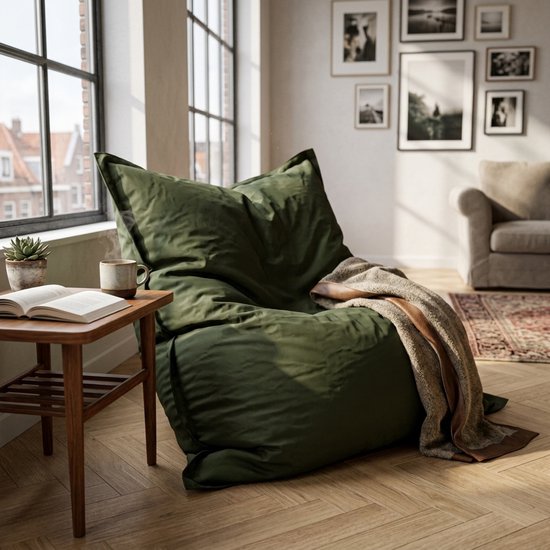 Grote donkergroene zitzak met een bruinwollige deken erop, in een woonkamer met grote ramen en zwart-wit foto's aan de muur.