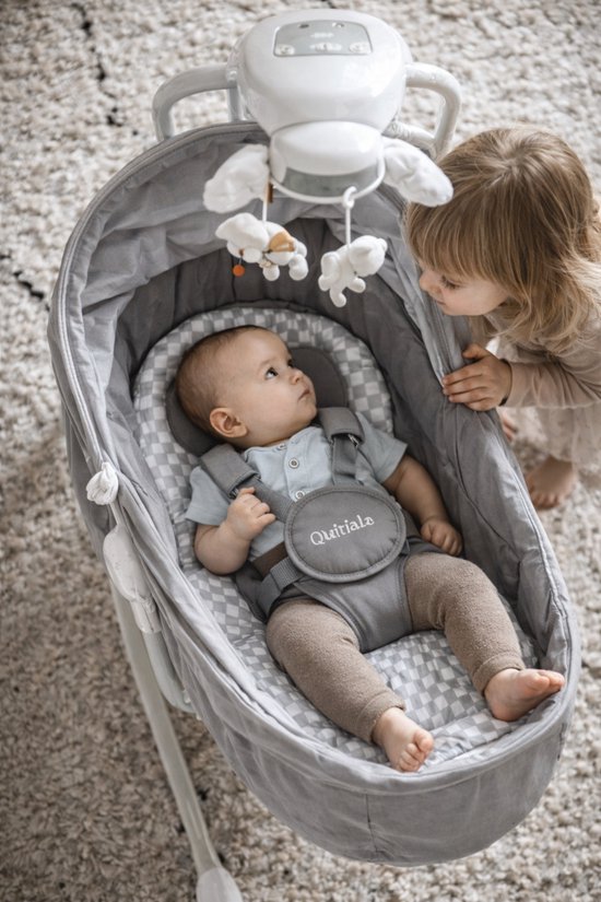 Baby in grijze wipstoel met speelgoedjes. Zus kijkt toe. Stoffering met ruitjes.