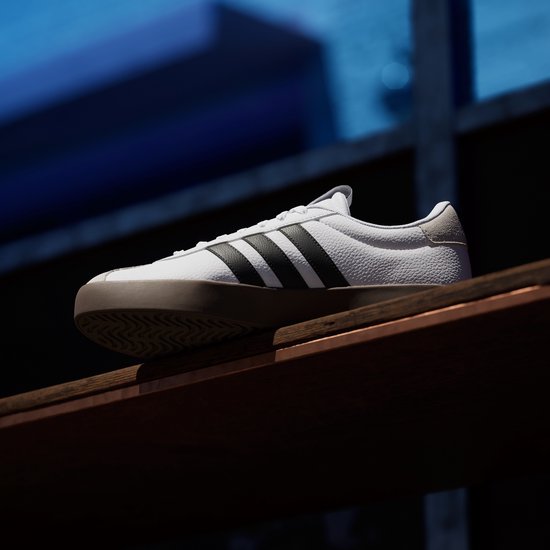 Wit leren Adidas VL Court 3.0 sneaker met bruine zool en zwarte strepen, geplaatst op een houten plank.