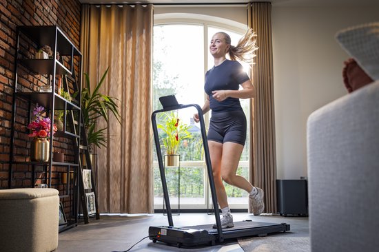 Vrouw loopt op een zwarte AJ-Sports loopband in een woonkamer, met zonlicht door het raam.