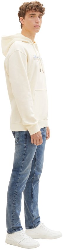 Man in crème hoodie en slanke, lichtblauwe spijkerbroek, gezien vanaf de zijkant.
