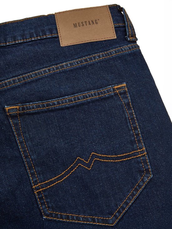 Close-up van de achterkant van een donkerblauwe denim jeans met oranje stiksels en een bruin Mustang label.