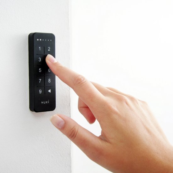 Vinger op de cijfertoets '2' van een zwart Nuki Keypad gemonteerd op een witte muur.