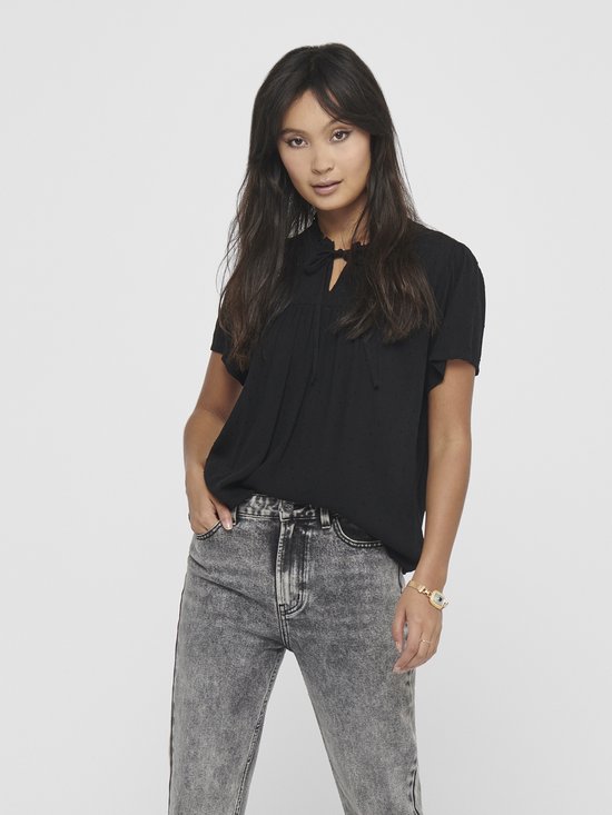 Model met zwarte blouse met korte mouwen, ruchekraag en strikdetail, gecombineerd met een grijze jeans.