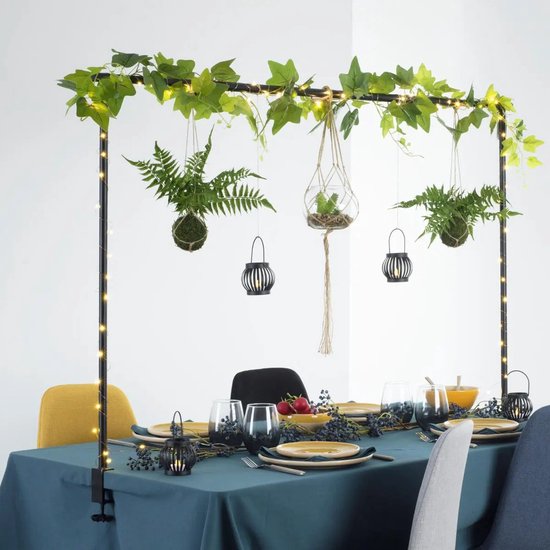 Zwarte metalen tafelbeugel met decoratieve planten en lampjes, boven een gedekte tafel.