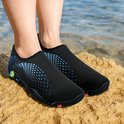 Voeten op een rots aan zee, dragend zwarte waterschoenen met blauwe spetters en kleurrijke zolen.