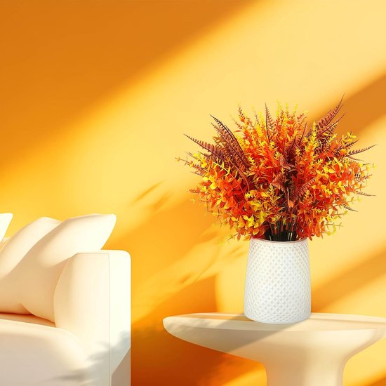 Plantes artificielles orange, jaune et brun rouille dans un vase blanc sur une table.