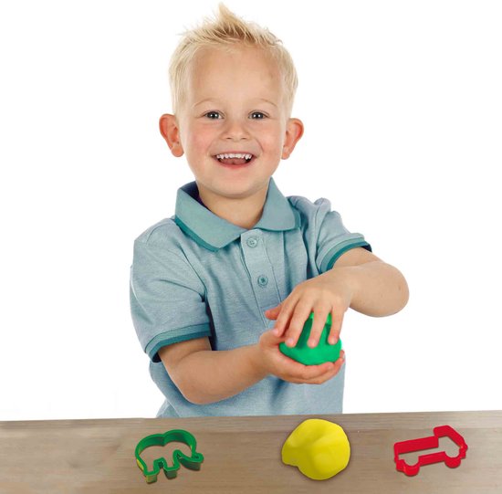 Een lachende jongen met blond haar houdt groen klei vast. Op de houten tafel liggen rode en groene uitsteekvormpjes.