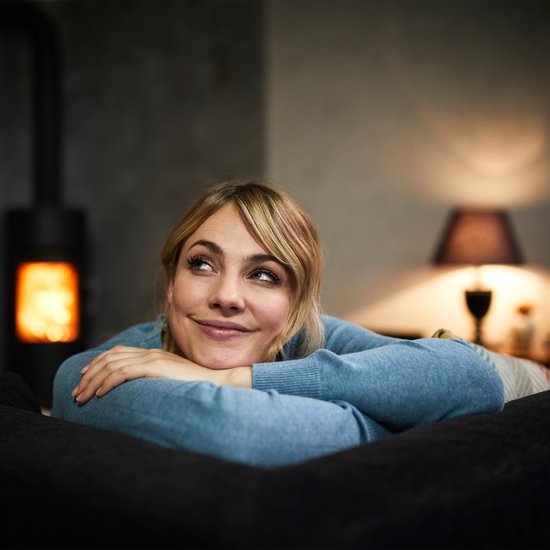 Vrouw met blond haar in blauwe trui ligt op een bank met op de achtergrond een kachel.