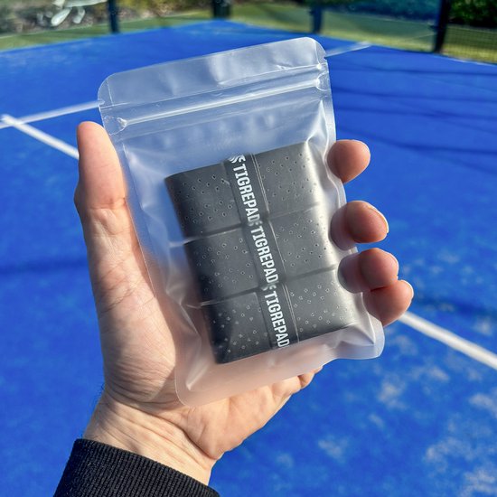 Hand houdt een doorzichtige verpakking vast met daarin vier zwarte TigrePadel overgrips met TigrePadel logo erop.