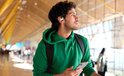 Man met krullend haar en groene hoodie draagt lichtblauwe JBL oordopjes tijdens het reizen.