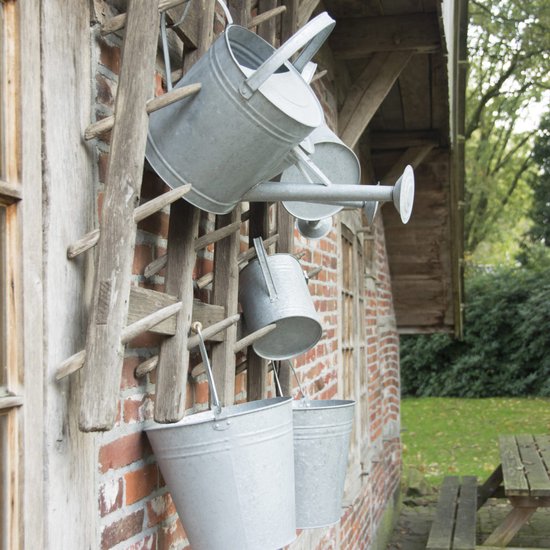 Drie zinken gieters en twee emmers hangen aan een houten rek tegen een bakstenen muur.