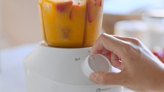 Hand draait aan de knop van een witte Philips blender, de blenderbeker bevat een oranje drank met stukjes fruit.