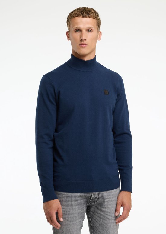 Man, vooraanzicht, draagt donkerblauwe coltrui en grijze jeans.