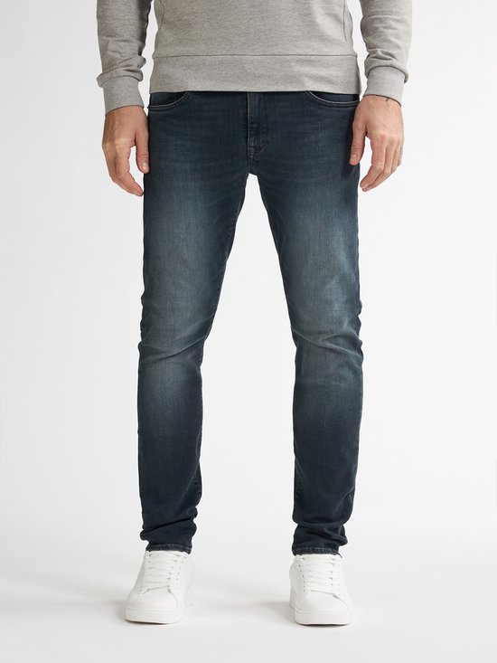 Man met donkerblauwe slim fit jeans, licht vervaagd aan de voorkant, met witte sneakers.