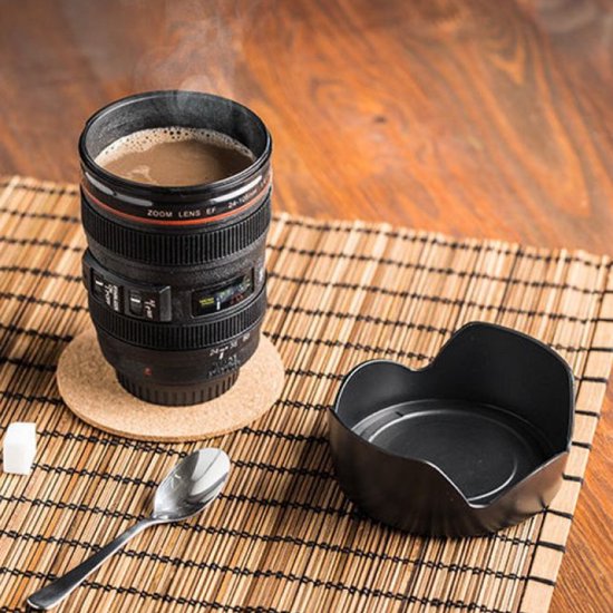Zwarte cameralens beker met warme koffie, staat op een bamboemat met een theelepeltje en suikerklontje.