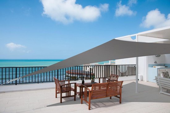 Antracietkleurig schaduwdoek driehoek 3x3m biedt schaduw op een terras met houten meubels aan zee.
