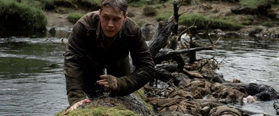 Een soldaat kruipt uit een rivier, nat en met modder besmeurd. Lijken drijven op de achtergrond.