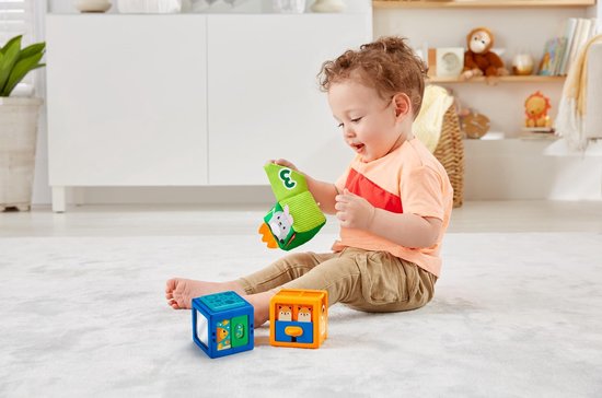 Baby speelt met Fisher-Price blokken op een kleed. Zacht konijnenblok met cijfer 3, blauw blok met cijfer 1, en oranje blok met vosjes.
