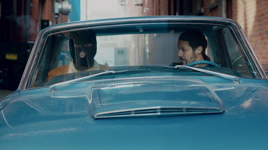 Twee mannen in een blauwe auto, één met zwart masker voor het gezicht, de ander kijkt opzij.