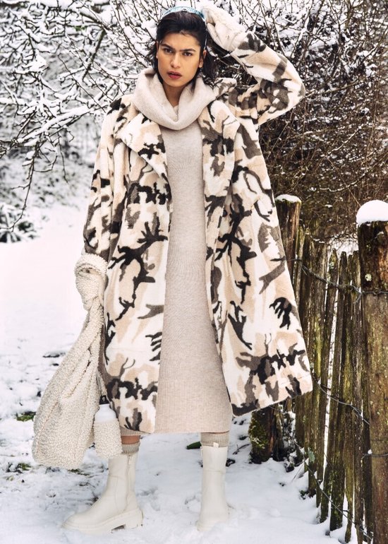 Mannequin portant un long gilet imprimé camouflage beige, marron et noir, dans la neige. Elle porte un col roulé tricoté et une robe.