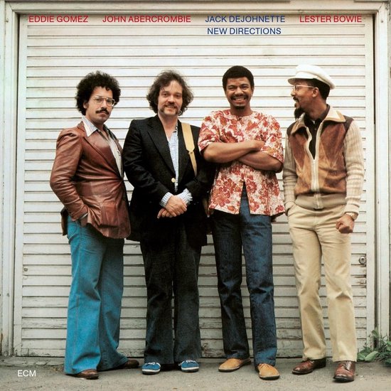 Vier mannen staan voor een garagedeur. Eddie Gomez draagt een bruine leren jas en blauwe broek. John Abercrombie een zwart pak. Jack DeJohnette een kleurrijk shirt en jeans. Lester Bowie een beige vest en broek.