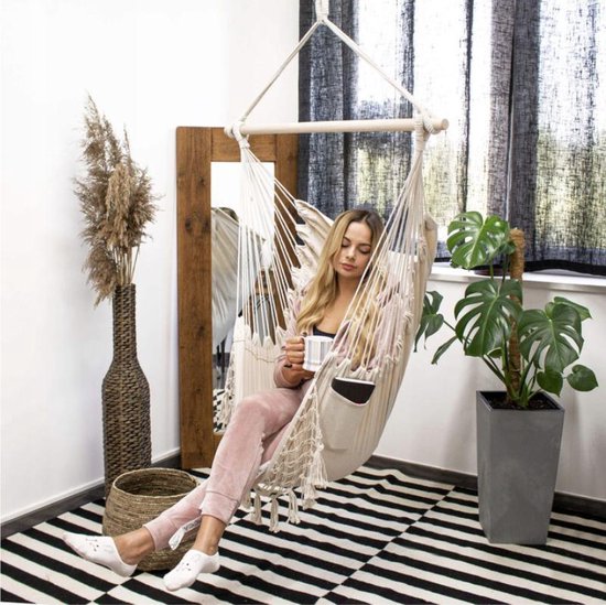 Vrouw met mok in lichte roze outfit zit in een witte hangstoel met macramé en franjes.