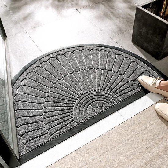 Halve cirkel donkergrijze deurmat met een reliëfpatroon van gestileerde bladeren of veren, en een zwarte rand. Gelegd op een lichte tegelvloer, met een beige schoen die erop stapt.