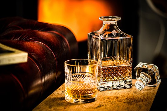 Een kristallen whiskykaraf en glas gevuld met amberkleurige drank, naast een leren bank voor een haardvuur.