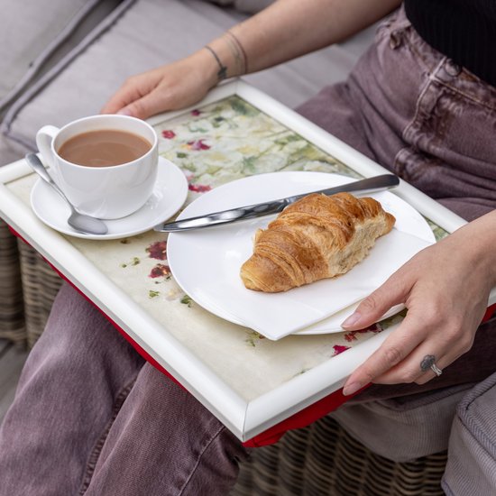 Vrouw met dienblad op schoot. Het dienblad heeft een bloemenprint van rode en witte klaprozen. Een kopje koffie en een croissant liggen erop.