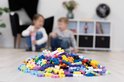 Een hoop veelkleurige Plus-Plus bouwstenen op de vloer met twee kinderen op de achtergrond.