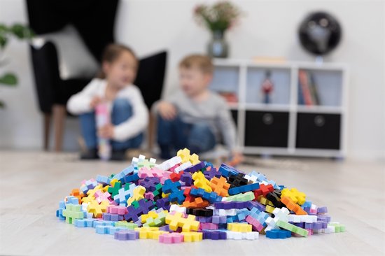 Een hoop veelkleurige Plus-Plus bouwstenen op de vloer met twee kinderen op de achtergrond.