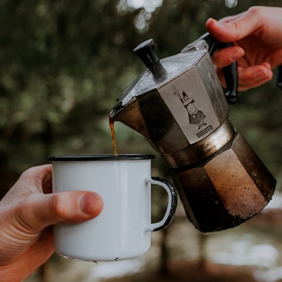 Gietend uit de Bialetti Moka Express espressopot in een wit emaille mok, buiten in het bos.