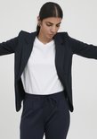 Model in een donkerblauwe blazer en een witte top, met wijd gespreide armen.