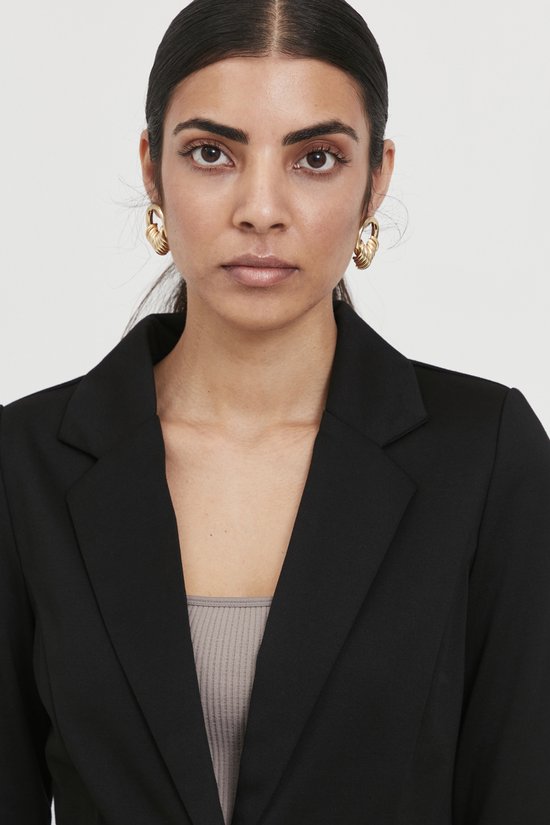 Vrouw met donker haar draagt een zwarte blazer over een beige top. Gouden oorbellen.