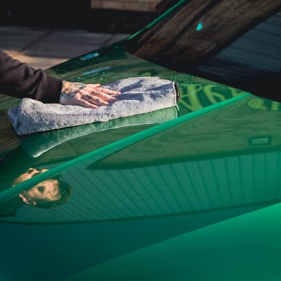 Hand met tatoeages droogt een glanzende groene auto met een grijze microvezel droogdoek.