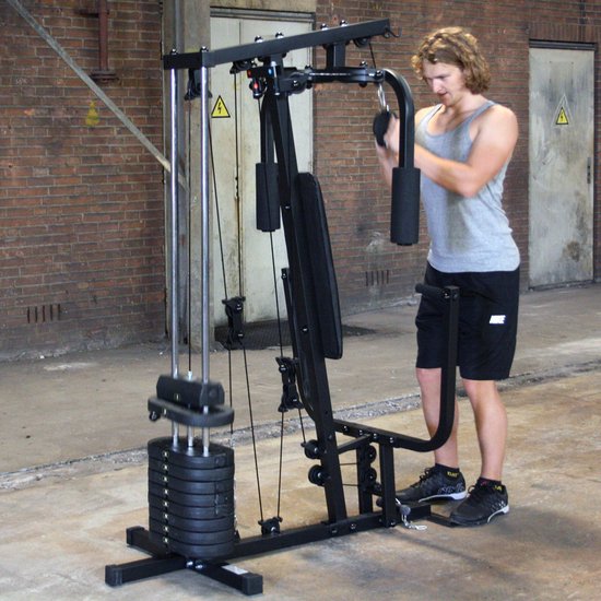 Man met krullend haar, grijs T-shirt en zwarte korte broek gebruikt de handgrepen van het Focus Fitness Unit 2 krachtstation.