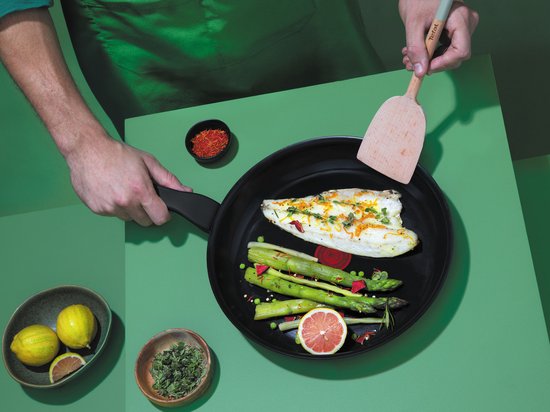Persoon bakt vis en asperges in een zwarte Tefal koekenpan op een groene ondergrond.