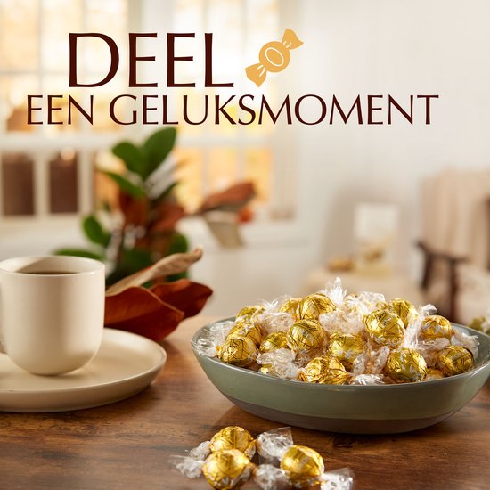 Een kom gevuld met Lindt LINDOR witte chocoladebonbons in goudfolie, naast een kop koffie.
