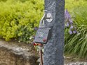 GARDENA besproeiingscomputer op stenen pilaar bij een kraan in de regen. Tuin met bloemen.
