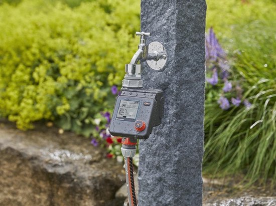 GARDENA besproeiingscomputer op stenen pilaar bij een kraan in de regen. Tuin met bloemen.