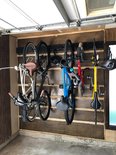 Zes fietsen, inclusief racefietsen en mountainbikes, hangen aan een zwart ophangsysteem in een garage.