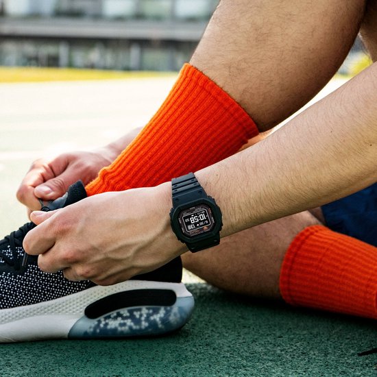 Zwart G-Shock horloge gedragen op de pols van een man met feloranje sokken, terwijl hij zijn schoen strikt.