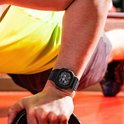Zwart G-Shock horloge om de pols van een man die een gewicht optilt, met een geel shirt op de achtergrond.