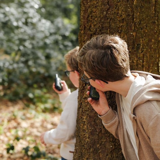 Twee jongens in een bos, achter een boom, die walkie talkies gebruiken.